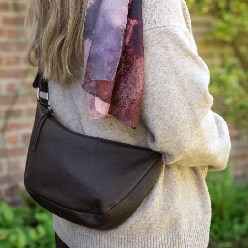 Black faux leather crossbody shoulder bag with a curved silhouette and adjustable strap, shown worn outdoors with a neutral knit jumper and patterned scarf to highlight the soft textured finish and everyday styling.