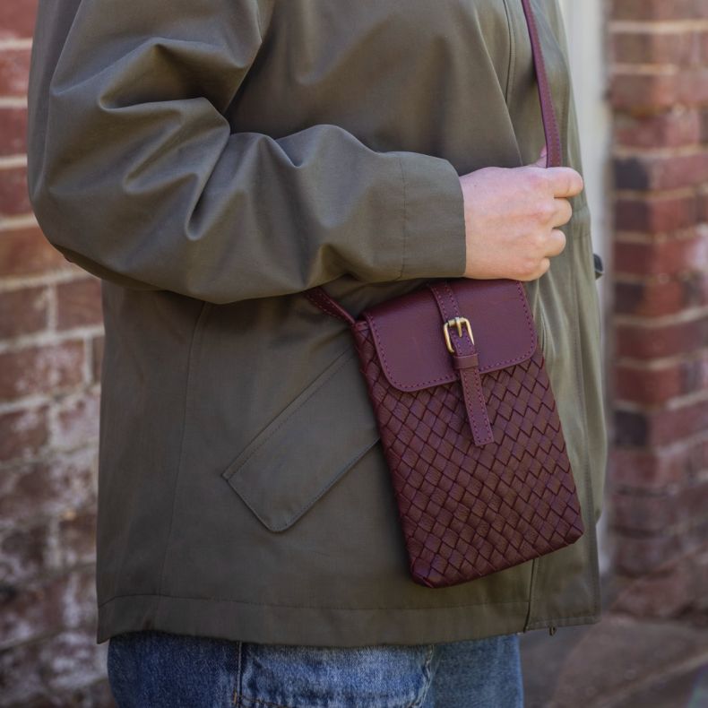Burgundy woven crossbody phone bag with a textured basketweave design, flap closure and adjustable strap, shown worn with an olive jacket to highlight the compact silhouette, detailed weave and everyday styling