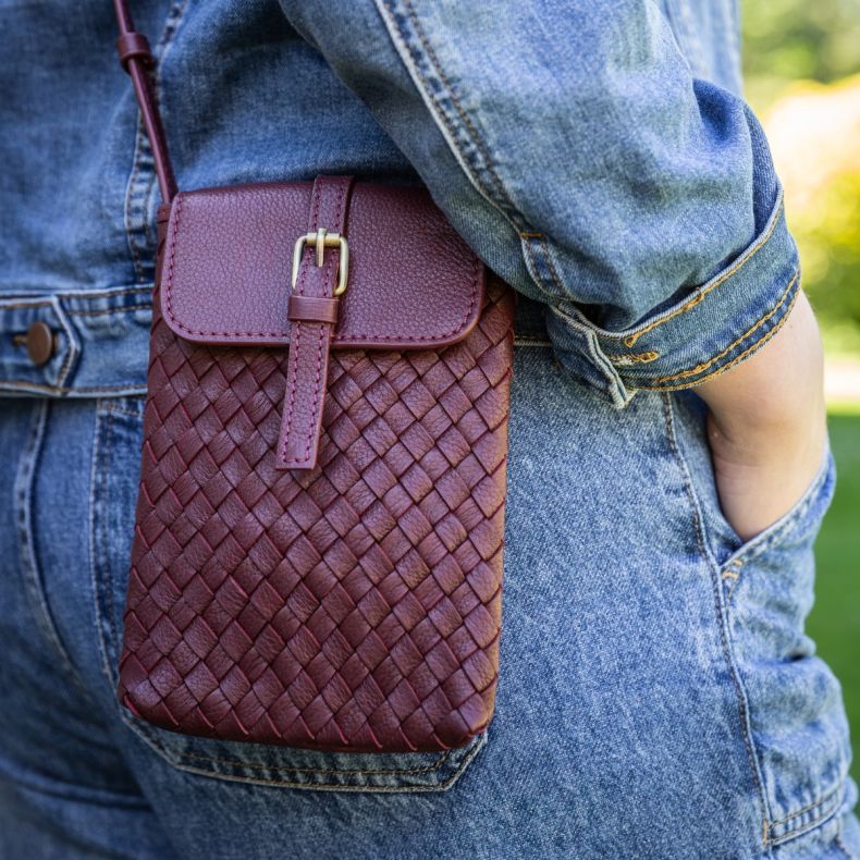 Burgundy woven crossbody phone bag with a textured basketweave design, flap closure and adjustable strap, shown worn with a denim jacket to highlight the detailed weave, compact shape and hands-free styling.
