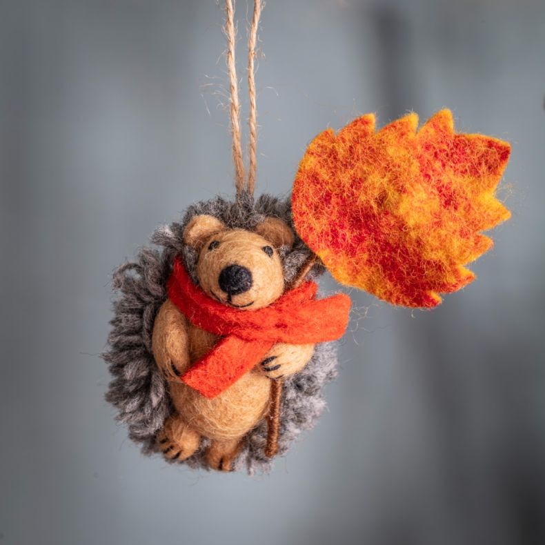 Felt hanging hedgehog ornament wearing an orange scarf and holding a vibrant orange autumn leaf, suspended by a natural hanging loop and designed as a seasonal woodland decoration.