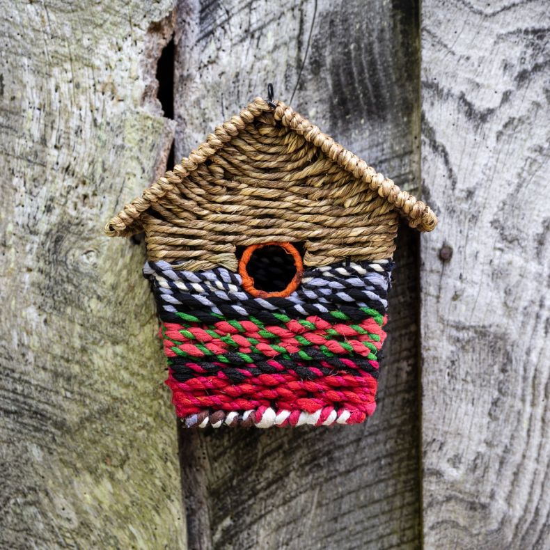 Handcrafted birdhouse ornament made from woven natural grass and recycled fabric, featuring a house-shaped design with a circular entrance hole, colourful braided textile detailing in pink, red, green, black and white tones, and a hanging loop for display