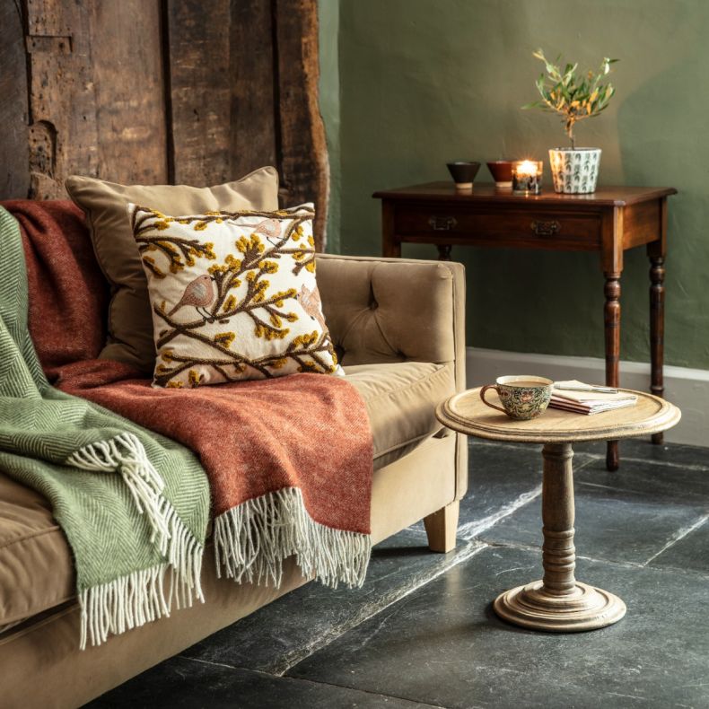 Embroidered decorative cushion featuring perched birds among textured yellow flowering branches on a cream background, styled on a sofa with green and rust woven throws in a cosy rustic living room setting.