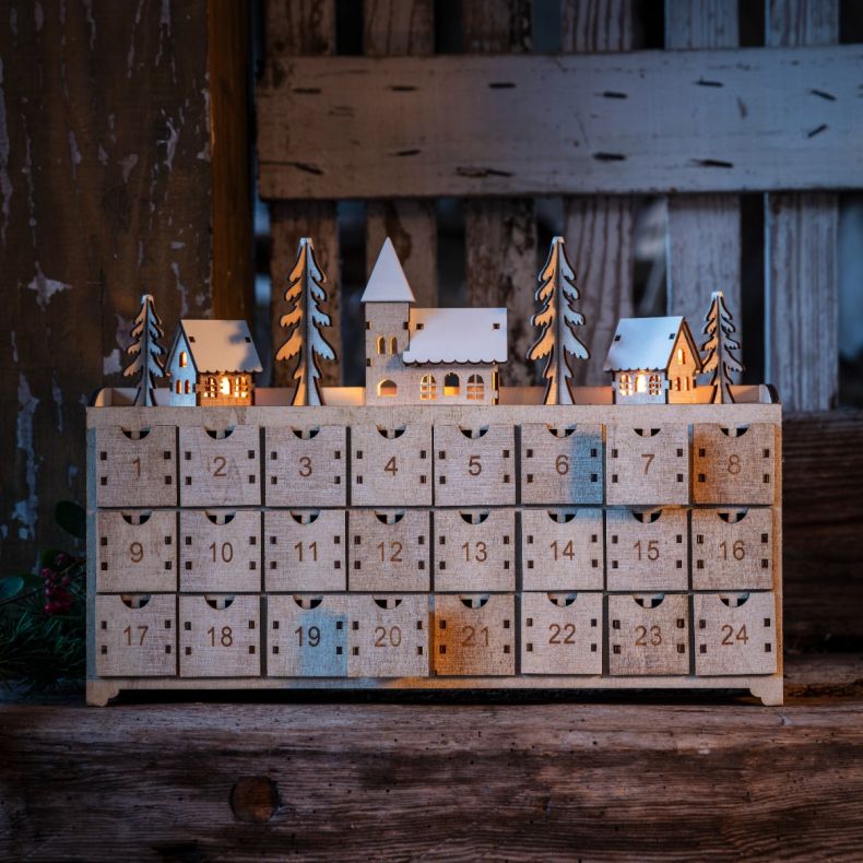 Wooden advent calendar featuring 24 numbered pull-out drawers beneath an illuminated winter village scene with snow-covered houses, church and fir trees, displayed in a rustic festive setting.