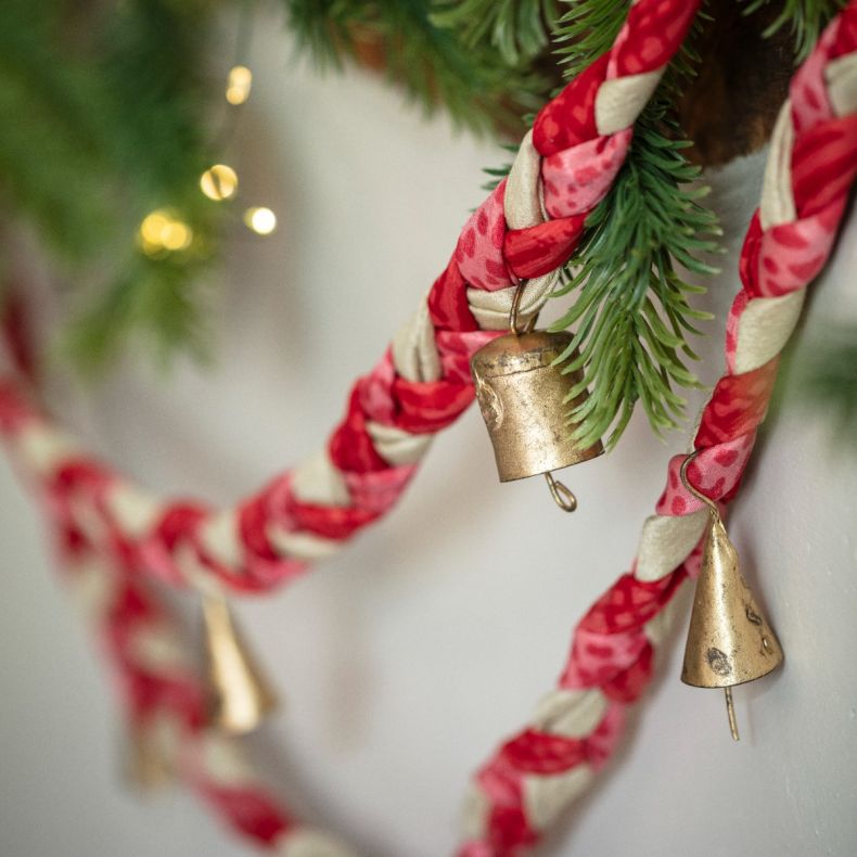 Close-up of a festive braided fabric garland in red, pink and cream tones featuring hanging brass bell decorations, displayed among evergreen foliage with warm fairy lights in the background.