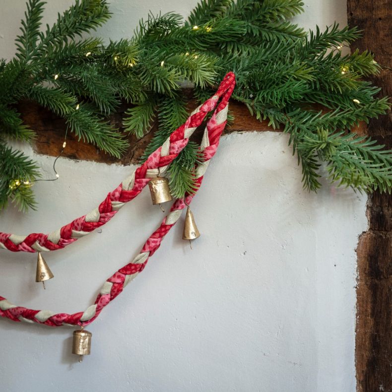 Festive braided fabric garland in red, pink and cream tones with hanging brass bell decorations, draped among evergreen foliage and warm fairy lights on a rustic mantel display.