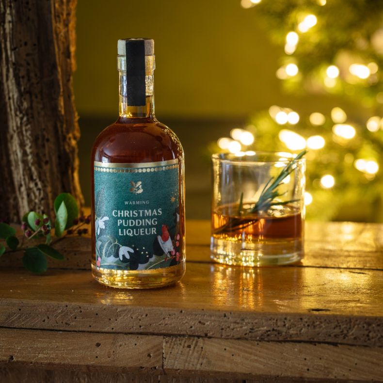 Christmas pudding liqueur in a clear glass bottle with wooden stopper, displayed alongside a glass of amber liqueur garnished with rosemary on a rustic wooden table, with warm festive lights in the background to showcase the seasonal spirit and serving su