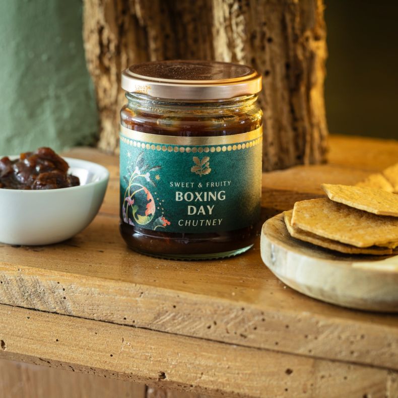 Boxing Day chutney in a glass jar with a silver screw-top lid, displayed on a rustic wooden table alongside crackers and a bowl of chutney to showcase the sweet, fruity flavour and serving suggestion.