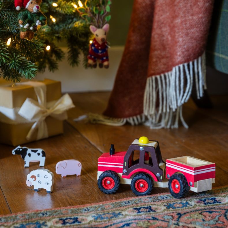 Wooden toy tractor with trailer in a red and natural wood finish, displayed beneath a decorated Christmas tree alongside wooden farm animal figures including a cow, pig and sheep, creating a festive farm-themed play scene.