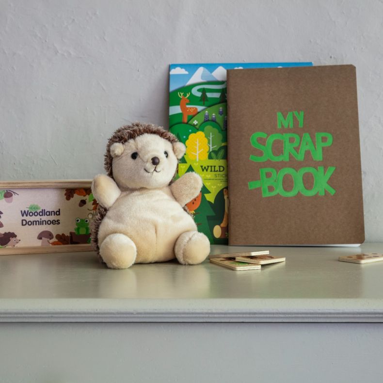 Soft plush hedgehog toy with a cream body and textured brown faux-fur spines, displayed alongside woodland-themed children's activities including a woodland dominoes set, nature sticker book and scrapbook on a nursery shelf.