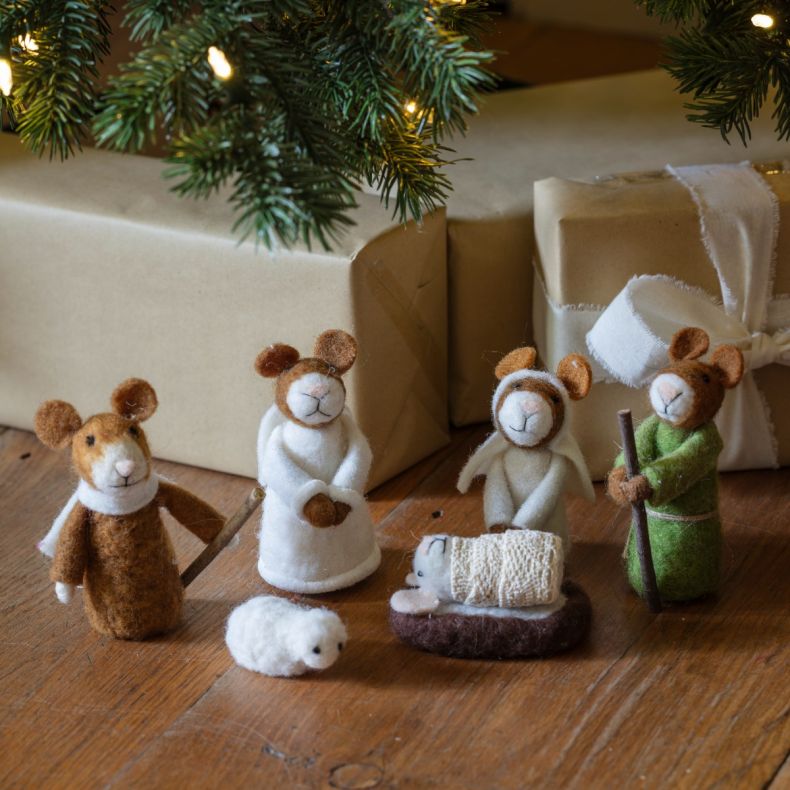 Felt nativity scene featuring mouse characters dressed as Mary, Joseph and an angel, with a swaddled baby in a manger and a small sheep figure, displayed beneath a decorated Christmas tree alongside wrapped festive gifts