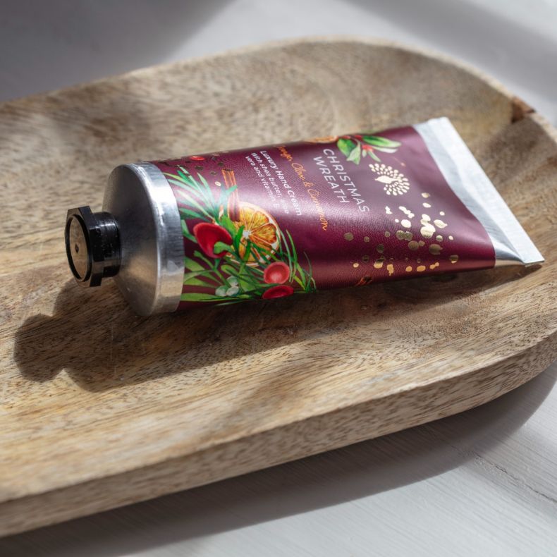 Christmas Wreath luxury hand cream in a decorative burgundy tube with festive botanical illustrations, displayed on a wooden tray to showcase the nourishing skincare product and seasonal gift-ready packaging.
