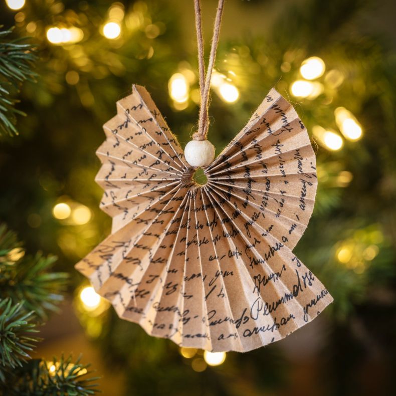 Folded paper angel hanging ornament crafted from brown paper printed with handwritten script, suspended by a natural hanging loop with a wooden bead detail and displayed against a Christmas tree with warm fairy lights in the background.