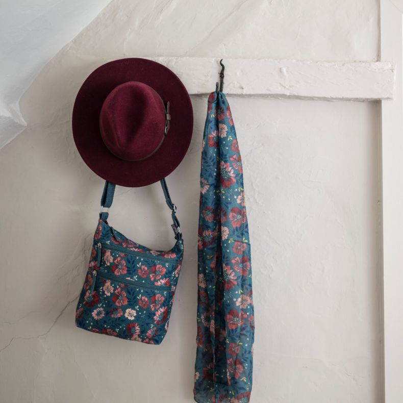 Blue floral print crossbody bag with adjustable shoulder strap and multiple zip compartments, displayed hanging beside a matching blue floral scarf and burgundy felt fedora hat to showcase the coordinated accessory collection.
