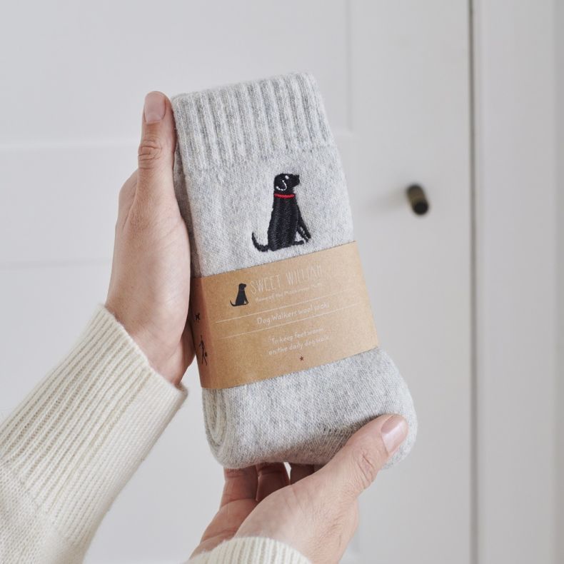 Sweet William wool socks for cat lovers in a soft beige marl knit, featuring an embroidered black labrador motif and presented with a branded kraft paper sleeve, shown held in two hands against a neutral background.