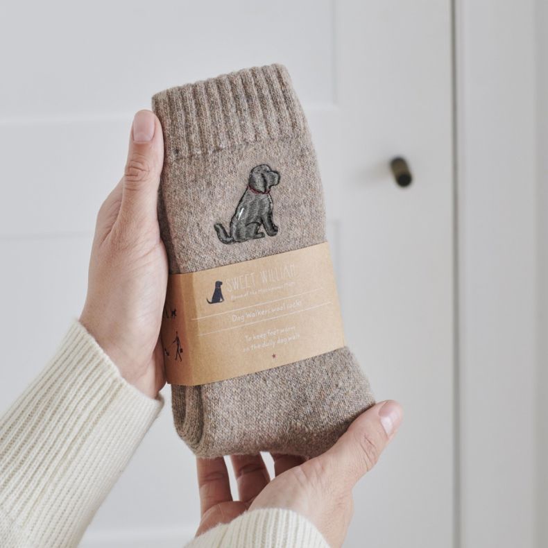Sweet William wool socks for cat lovers in a soft beige marl knit, featuring an embroidered grey cockapoo motif and presented with a branded kraft paper sleeve, shown held in two hands against a neutral background.