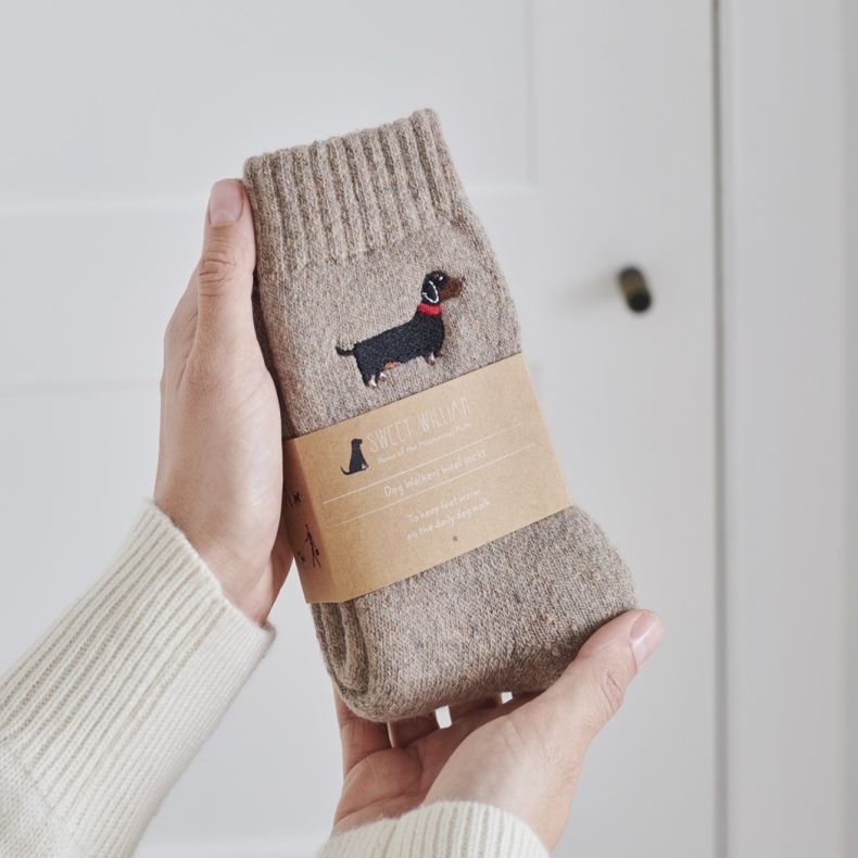 Sweet William wool socks for cat lovers in a soft beige marl knit, featuring an embroidered brown daschund motif and presented with a branded kraft paper sleeve, shown held in two hands against a neutral background.