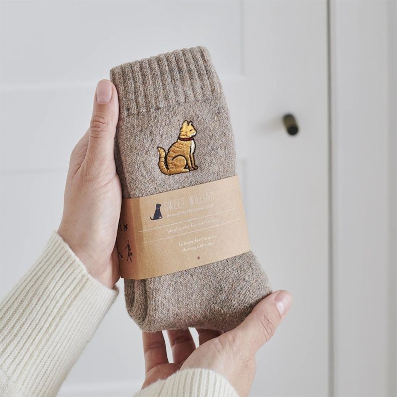 Sweet William wool socks for cat lovers in a soft beige marl knit, featuring an embroidered ginger cat motif and presented with a branded kraft paper sleeve, shown held in two hands against a neutral background.