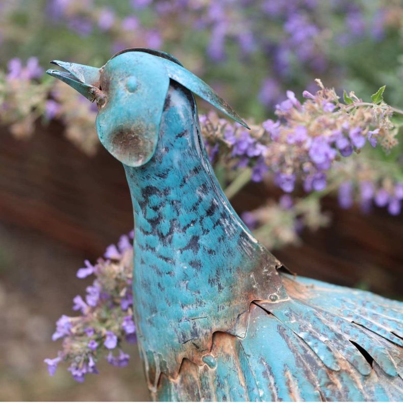 Close-up of a hand-painted antique metal pheasant sculpture with a verdigris finish, showcasing its detailed head and neck against a backdrop of purple lavender flowers.