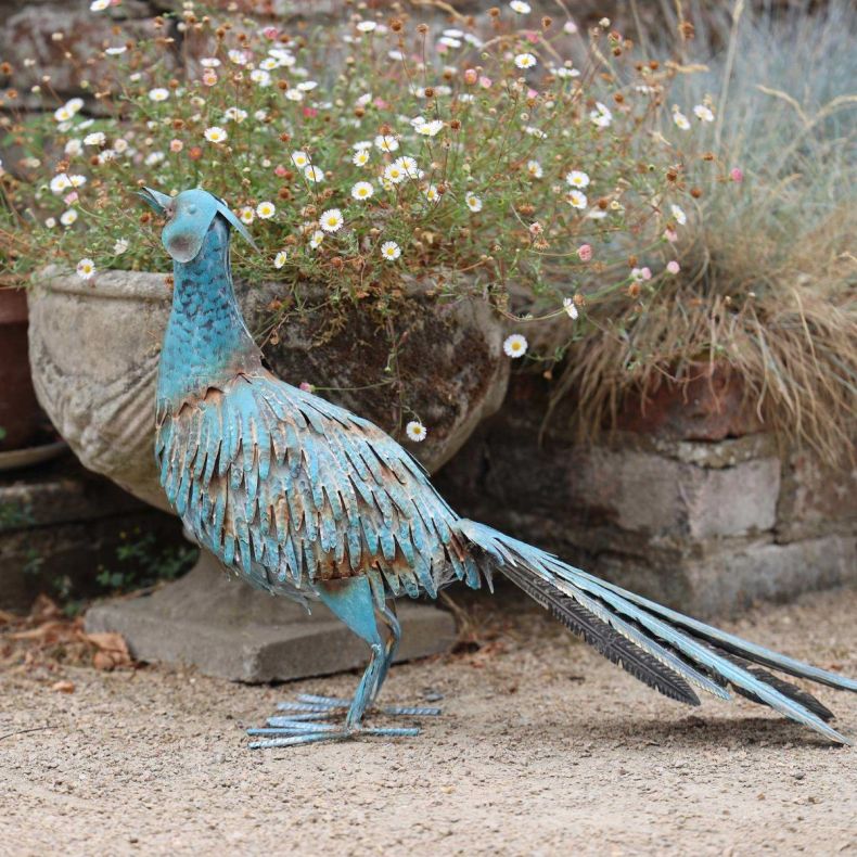 Hand-painted antique metal pheasant garden sculpture with a verdigris finish standing on a gravel path beside a stone planter filled with flowers.