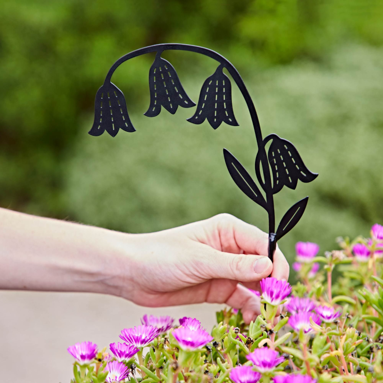 Recycled Metal Bluebell Silhouette Stake featuring four bell-shaped bluebell flowers and leaf details cut from black recycled metal, shown being placed into a flower bed as a decorative garden border or planter accent.