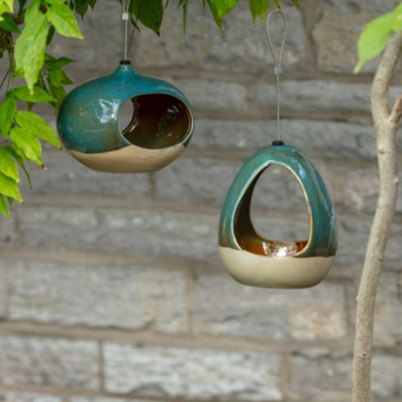 Two ceramic bird feeders in glazed green and cream tones hanging from tree branches. One has a round pod shape, and the other a teardrop design, both suspended in front of a stone wall with leafy branches above.