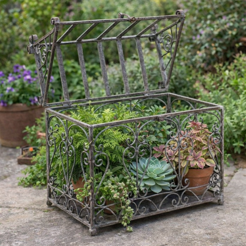 Victorian-style metal plant cloche with ornate scrollwork, filled with ferns and succulents, displayed on a stone surface in a garden setting.
