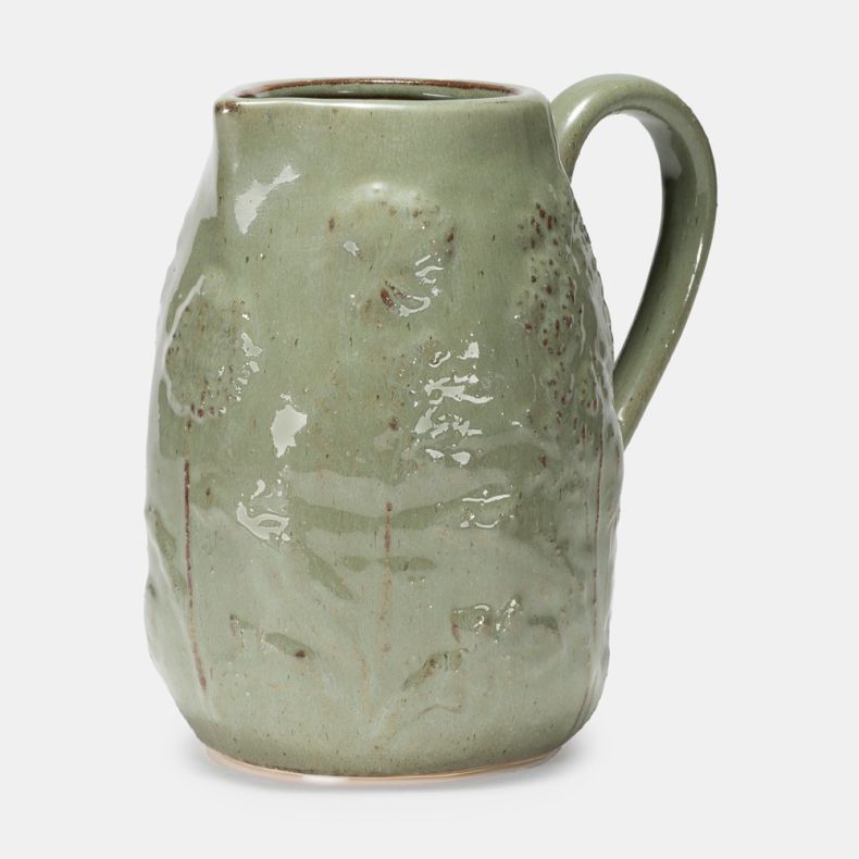Sage green ceramic jug with a glossy glazed finish and embossed meadow-inspired floral detailing. Shown from a front angled view, the jug features a curved handle, subtle speckled texture and handcrafted appearance against a plain light grey background.