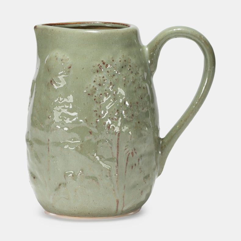 Sage green ceramic jug with a glossy glazed finish and embossed meadow-inspired floral design. The handcrafted-style pitcher features a curved handle, pouring spout and subtle speckled detailing, shown against a plain light grey background.