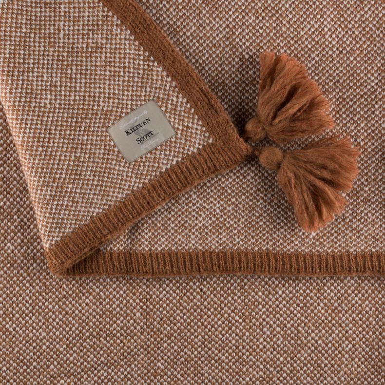 Close-up of a woven beige and cream throw blanket featuring a textured knit pattern, brown ribbed edging and decorative tassels. A branded fabric label is visible on the folded corner, highlighting the blanket’s craftsmanship and detail.