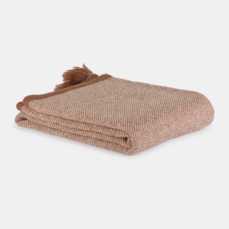 Folded woven throw blanket in a soft beige and cream pattern, neatly stacked with fringed tassels visible at one end. The textured fabric is shown against a plain light grey background.