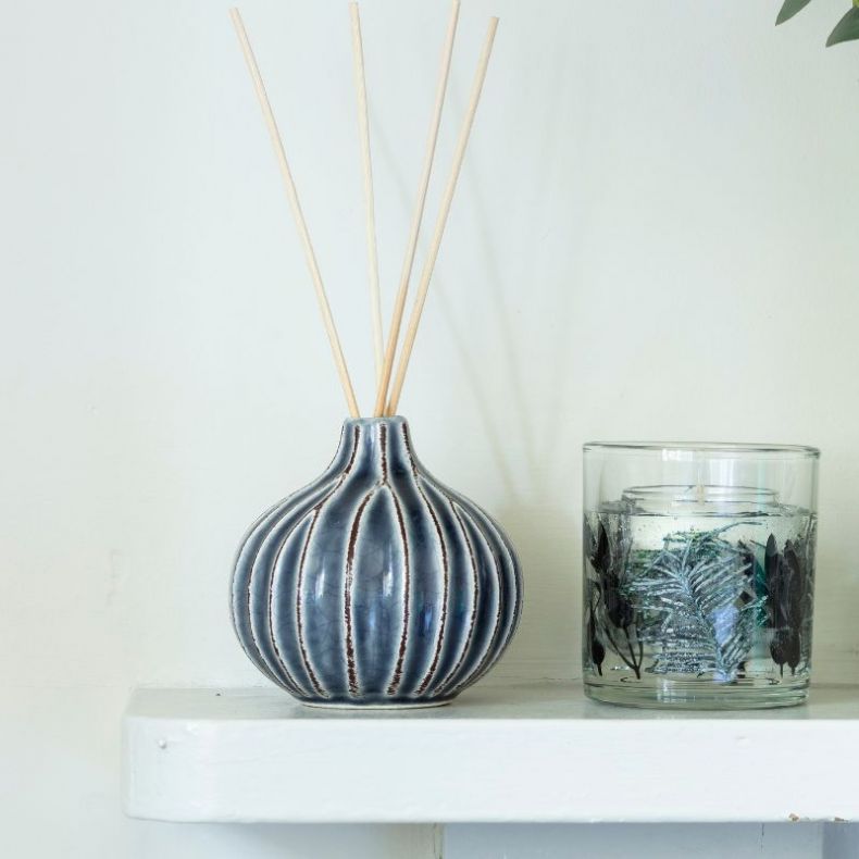 Blue-grey ceramic reed diffuser with a rounded, ribbed silhouette and narrow neck, featuring a glossy crackle-effect glaze and natural reed sticks extending from the top. The diffuser is displayed on a white shelf beside a decorative gel candle in a clear