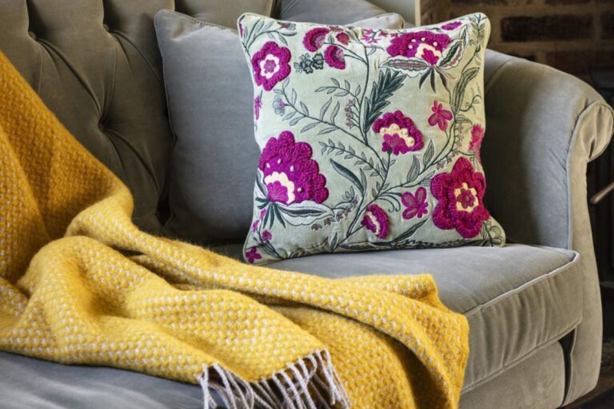 Cosy autumn winter living room scene featuring a grey sofa with a floral embroidered cushion, a yellow throw, and a log burner glowing in the background.