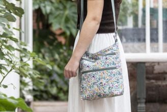 Bags - Woman wearing a National Trust crossbody bag with a colourful floral print, standing in a bright, leafy setting.
