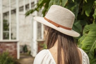 Hats - Woman wearing a straw sun hat with a tan band, standing near a greenhouse surrounded by greenery.