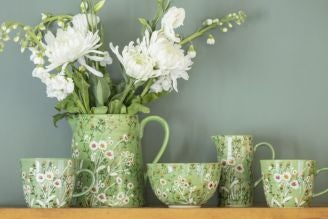 A green floral ceramic tableware set displayed on a wooden shelf against a muted green wall. The set includes a large jug filled with white flowers, two mugs, a bowl, a small jug, and an espresso cup, all decorated with delicate white and red flowers and 