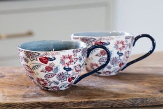 A green floral ceramic tableware set displayed on a wooden shelf against a muted green wall. The set includes a large jug filled with white flowers, two mugs, a bowl, a small jug, and an espresso cup, all decorated with delicate white and red flowers and 