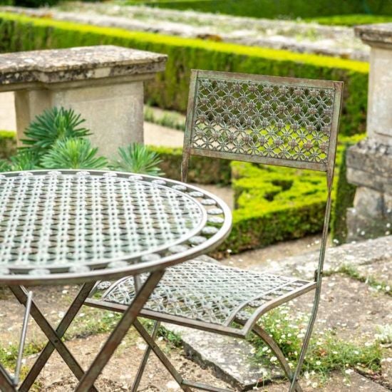 Vintage-style metal bistro table and two chairs set in a formal garden with stone balustrade and manicured hedges..