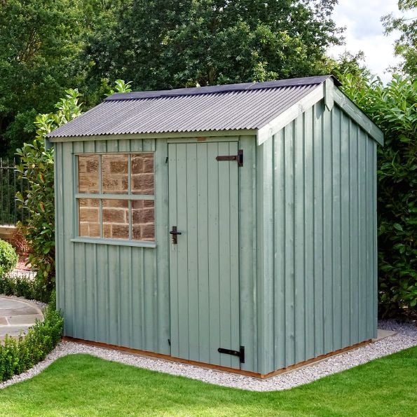 A small sage green wooden garden shed with vertical cladding and a dark corrugated roof, set on a gravel base in a neatly kept lawn. The shed has a single plank door with black hinges and latch, and a square window with wooden framing and multiple panes o