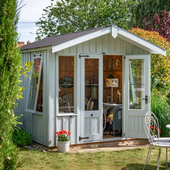 A small pale grey wooden garden room with a pitched roof and white trim, set on a lawn surrounded by greenery. The front features glazed double doors and tall side windows, revealing a cosy interior with wooden walls, a desk, chair and table lamp. A potte